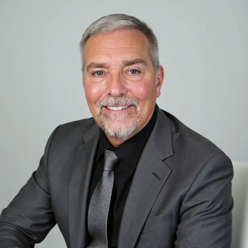 Smiling middle-aged man with short gray hair and beard, wearing a gray suit, black shirt, and gray tie, seated on a white chair against a plain light-gray background.