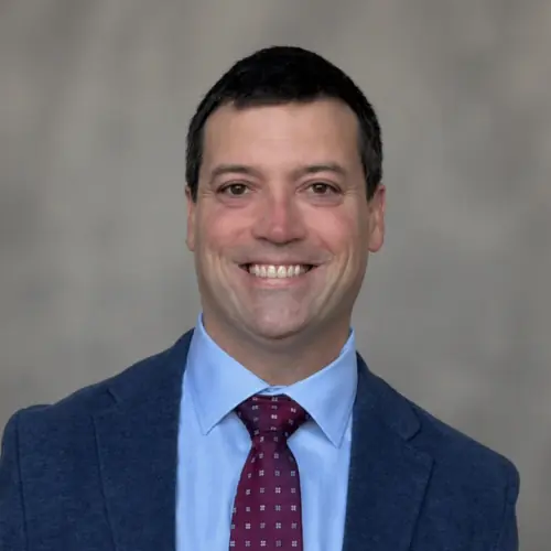 A man in a blue suit, light blue shirt, and maroon patterned tie smiles at the camera against a plain, neutral background.
