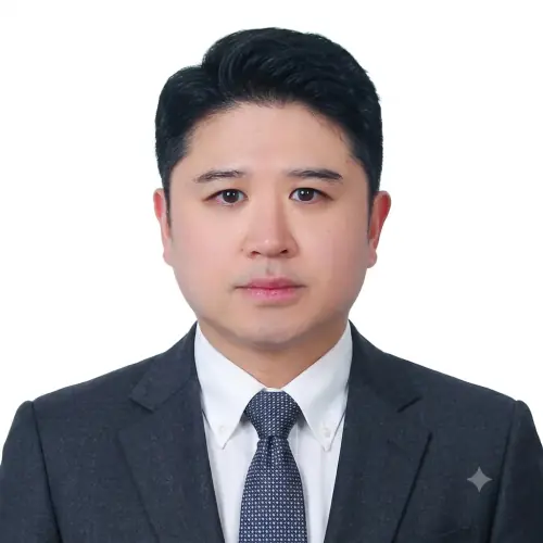 A man with short black hair wearing a dark suit, white shirt, and patterned tie poses for a formal headshot against a plain white background.